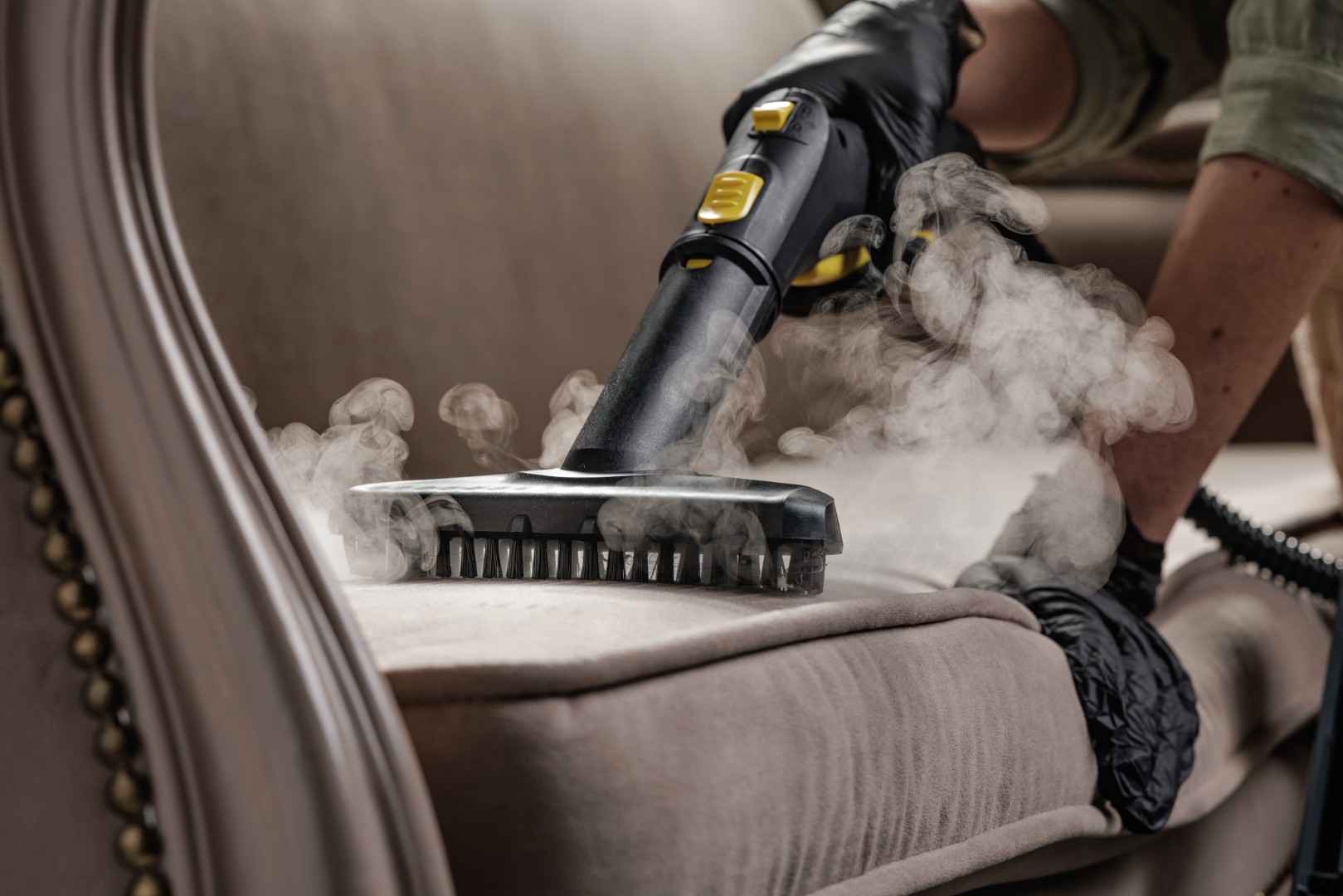 Close view of steam brush with vapor treating sofa cushion for deep upholstery sanitation and home hygiene service